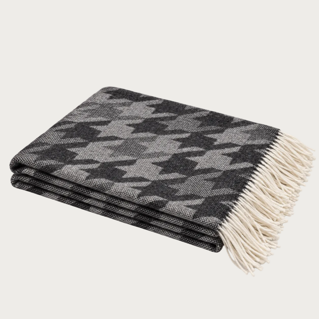 A folded woven throw blanket with a black and gray houndstooth pattern and cream fringe, photographed on a light warm gray background.