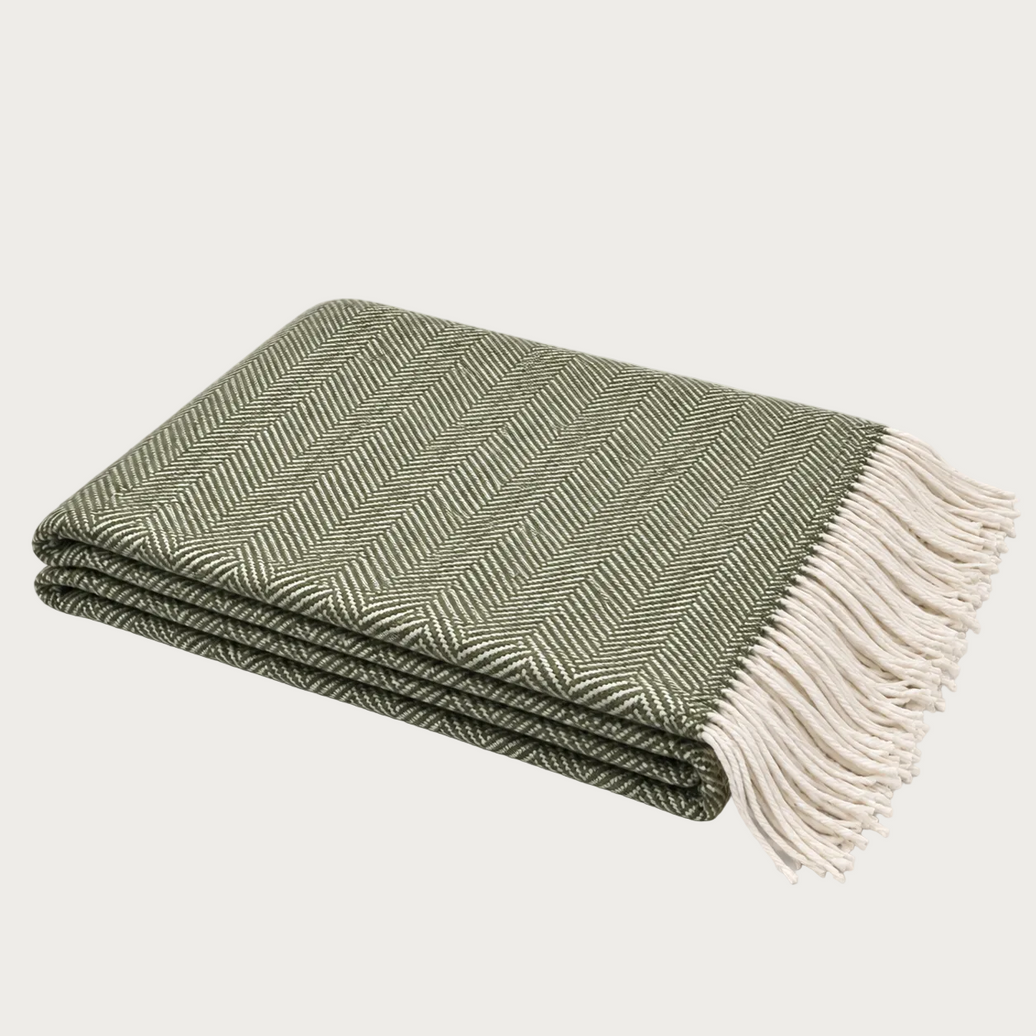 A folded woven throw blanket with a green and off-white herringbone pattern and cream fringe, photographed on a light neutral background.