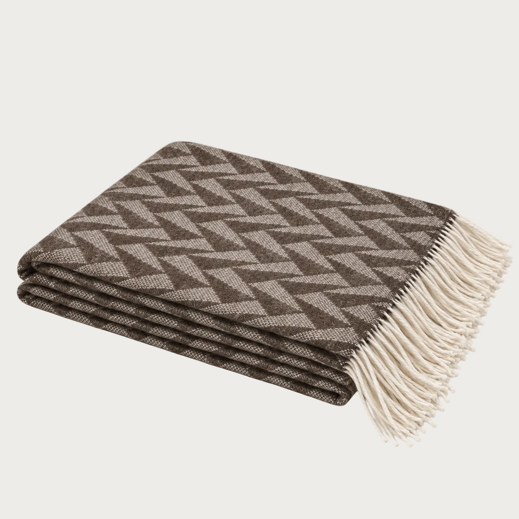 A folded woven throw blanket with a brown and beige geometric herringbone pattern and cream fringe, photographed on a clean off-white background.