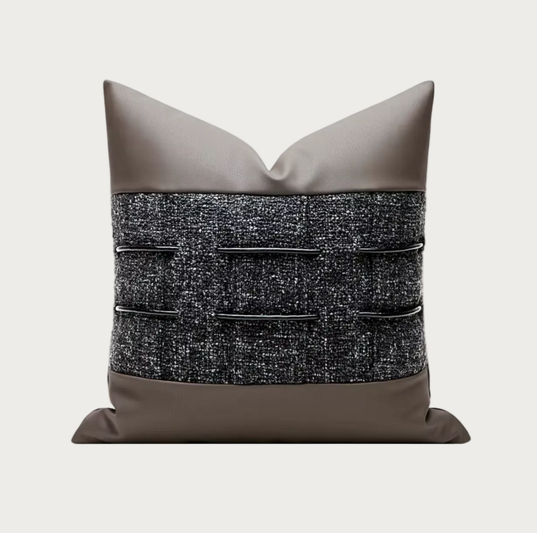 A square decorative throw pillow with a taupe faux-leather top and bottom and a textured black and gray tweed center panel featuring horizontal black leather tabs.