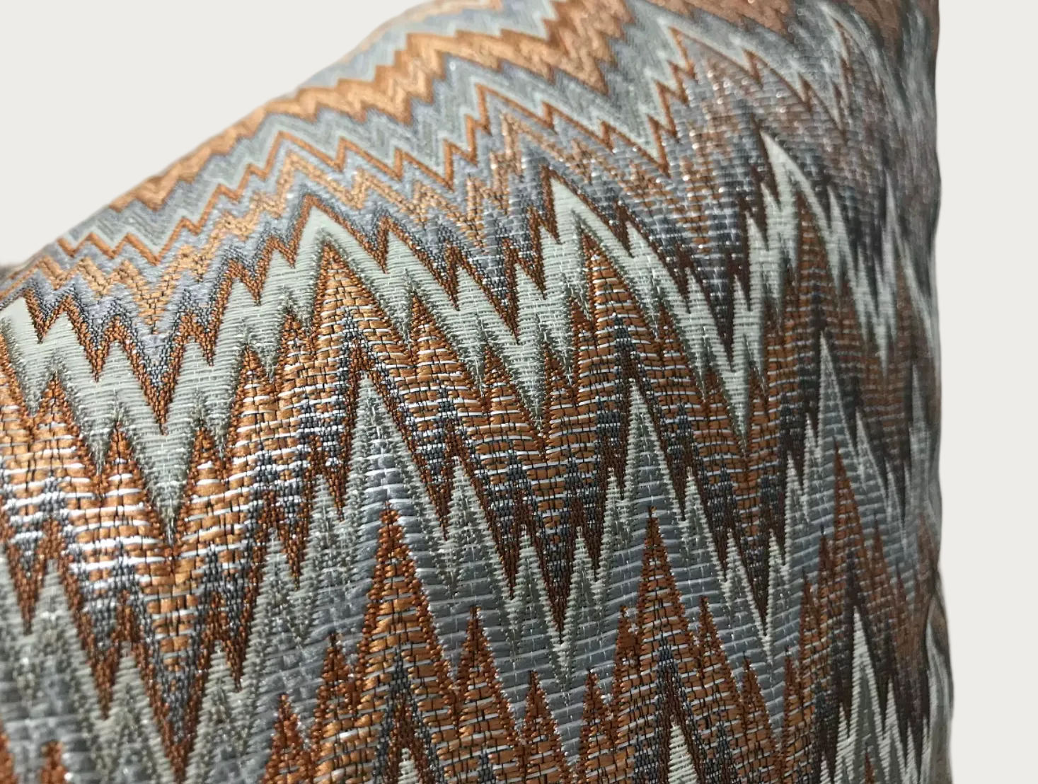 Close-up of a woven throw pillow cover featuring a geometric zigzag pattern in metallic bronze, gray, and off-white.
