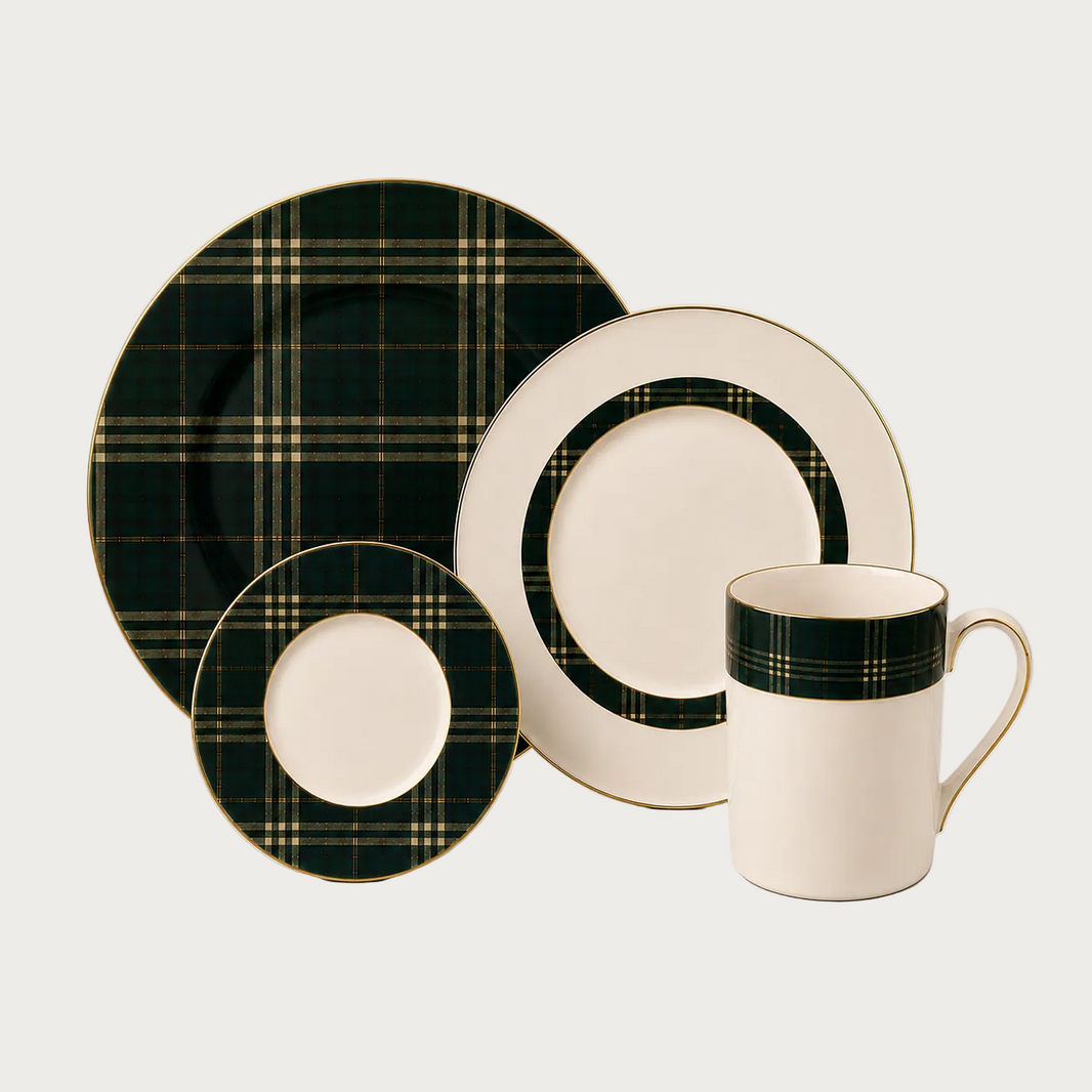 A dinnerware set on a light warm-gray background, featuring a large plate, a smaller plate, a saucer, and a mug. The plates and mug are cream-colored with a dark green and gold plaid pattern around the rims.