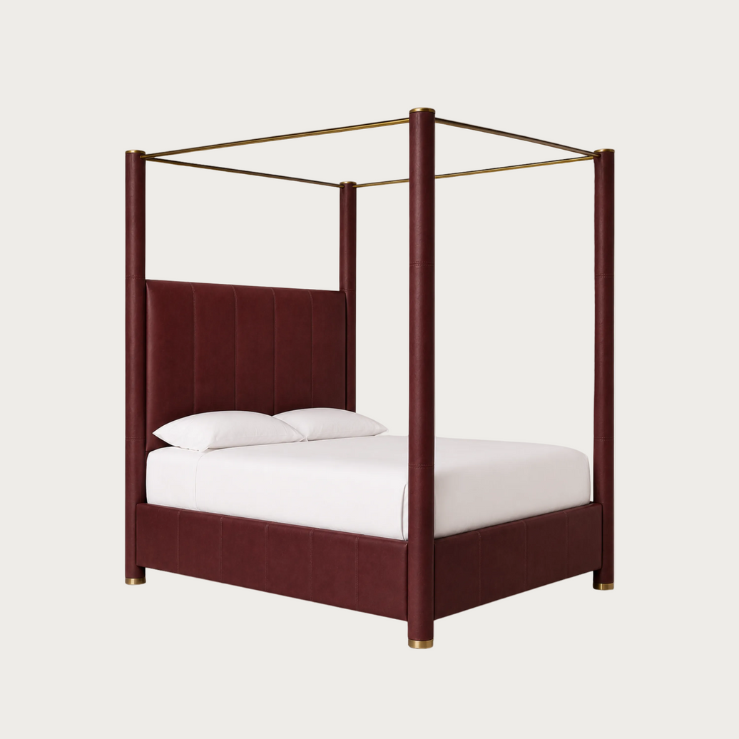 A luxury canopy bed featuring deep burgundy tufted upholstery on the headboard and base, supported by four vertical posts with gold-finished caps, and topped with a gold brass canopy frame. The bed is styled with white bedding and pillows against a solid off-white background.