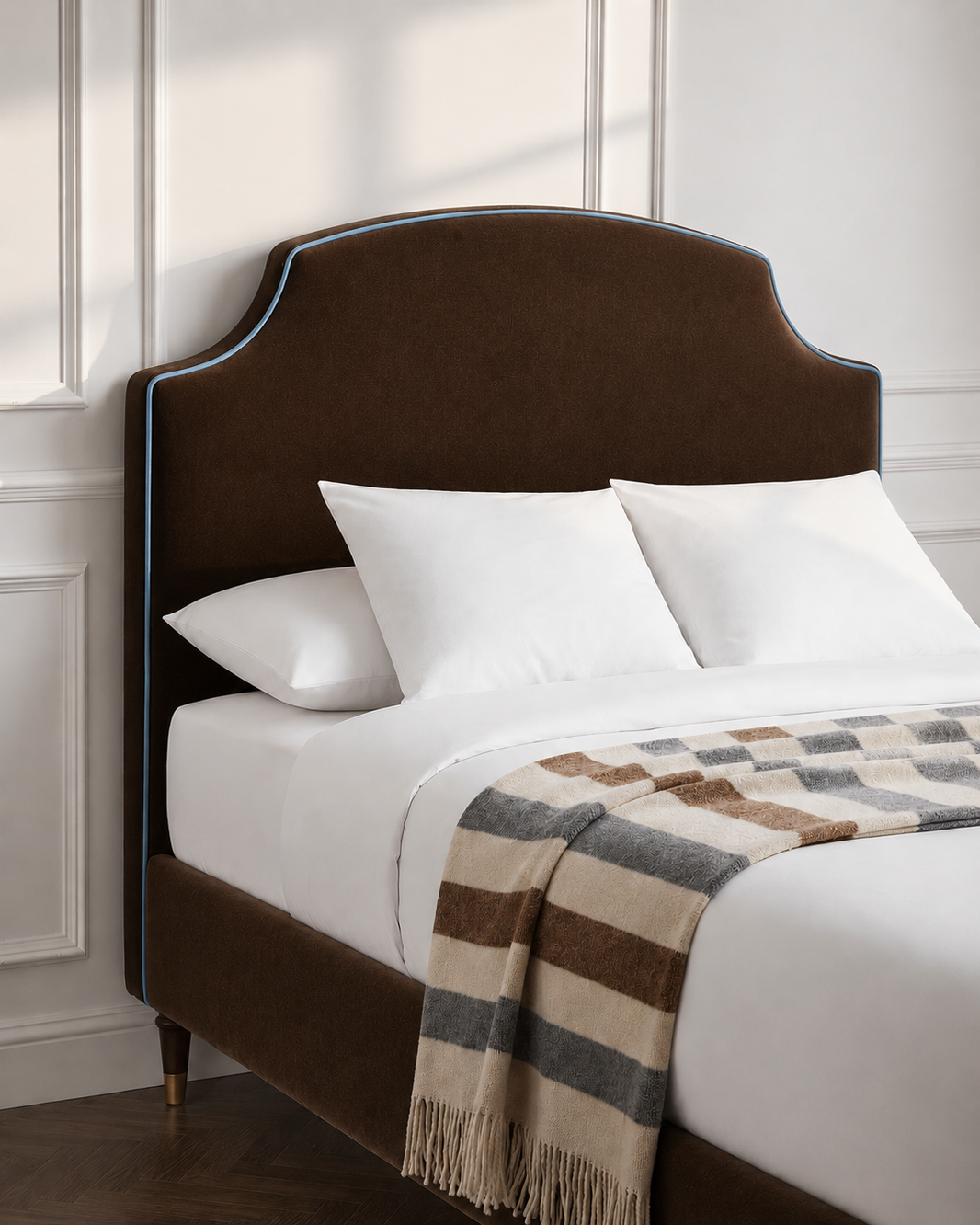 A dark brown upholstered bed frame with a curved headboard and matching footboard, styled with white bedding and a striped brown, beige, and gray throw blanket against a white paneled wall.